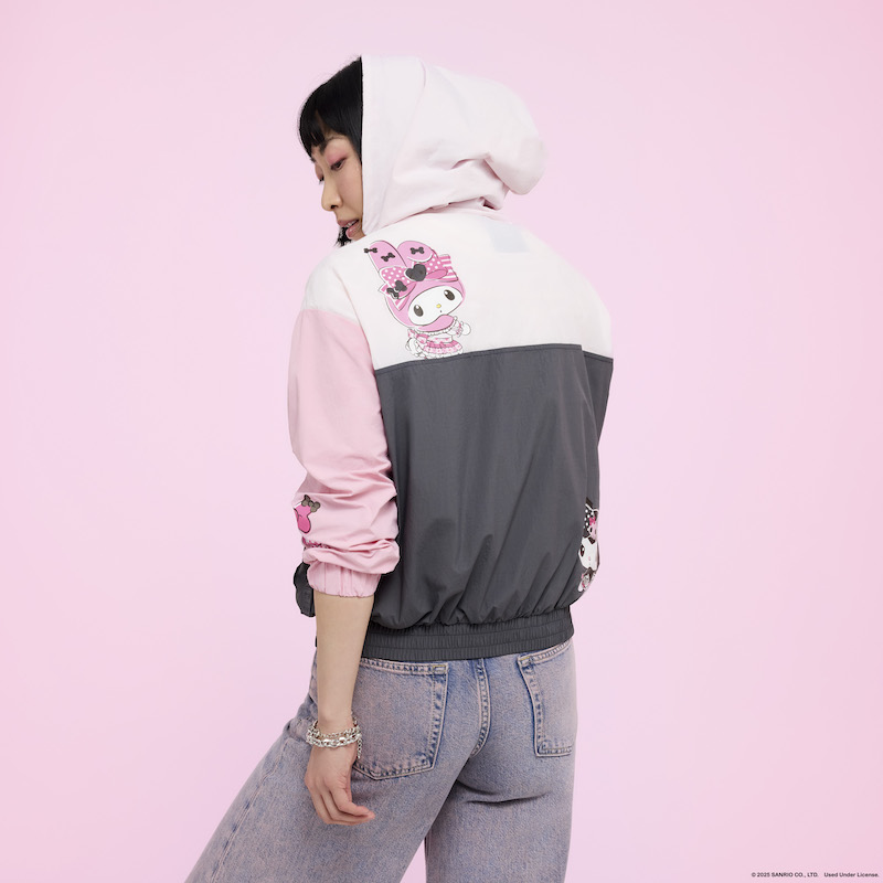 Woman facing away from camera wearing the Loungefly Sanrio My Melody & Kuromi Unisex Windbreaker Jacket with the hood up. The back of the jacket features My Melody in the upper corner against a pink background and Kuromi in the lower corner against a black background. 