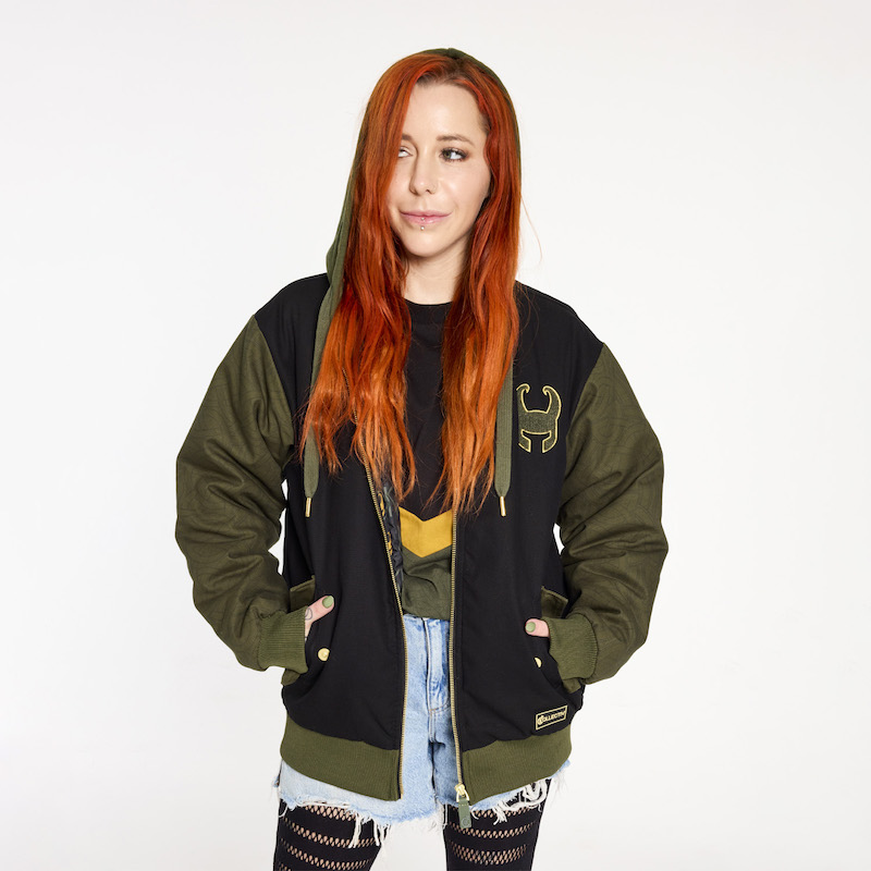 Redheaded woman wearing the Loungefly COLLECTIV Marvel Loki The WEEKENDR Hooded Jacket, with the hood up, over the Marvel Loki The ORIGINL Tee, with light blue denim shorts and ripped black leggings. 