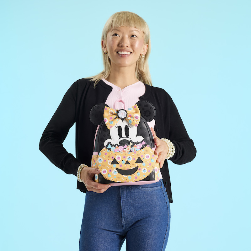 Woman wearing a pink top under a black sweater with blue jeans. She holds the Loungefly Minnie Mouse Cosplay Floral Pumpkin Glow mini backpack. She stands in front of a blue background. 