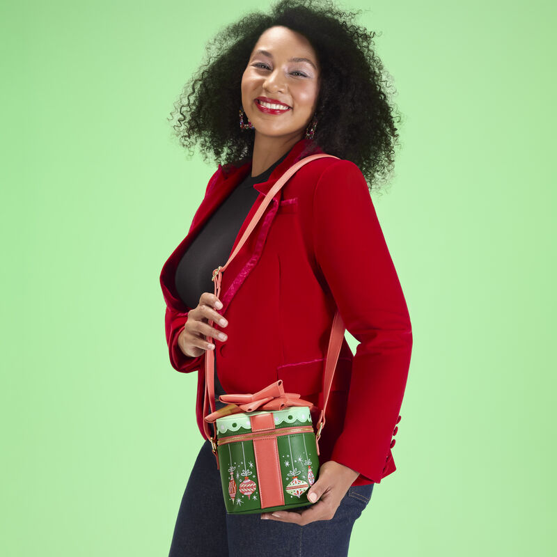 Woman wearing a black top and a red jacket, wearing the Loungefly holiday gift figural crossbody bag. She stands against a green background. 