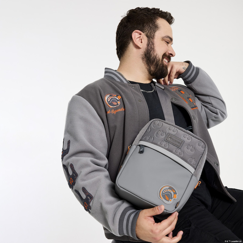 Bearded man sitting and looking over his left shoulder wearing the Loungefly COLLECTIV Star Wars Rebel Alliance VRSITY Jacket and wearing the Loungefly COLLECTIV Star Wars Rebel Alliance The EVRYDAY Convertible Bag as a sling, holding the bottom edge of the bag with his right hand