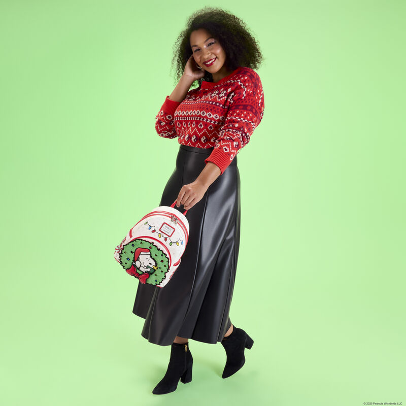 Woman wearing a patterned red sweater and long, black leather skirt. She holds the Loungefly Peanuts holiday wreath mini backpack. She stands against a green background. 