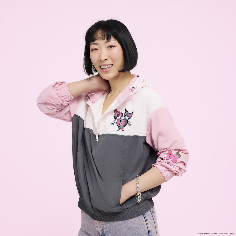 Woman posing in front of camera wearing the Loungefly Sanrio My Melody & Kuromi Unisex Windbreaker Jacket, featuring an image of My Melody & Kuromi over a heart in the upper right corner. She stands against a light pink background. 
