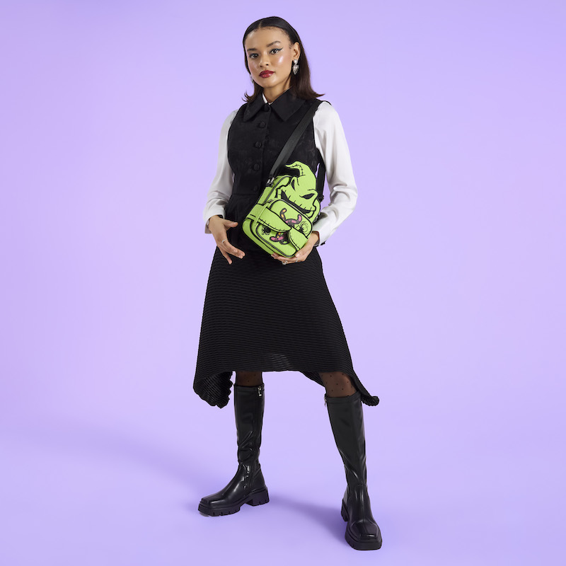 Woman wearing a white long-sleeve top under a black dress. She wears the Loungefly Nightmare Before Christmas Oogie Boogie Crossbuddies Crossbody bag over a shoulder. She stands in front of a purple background. 