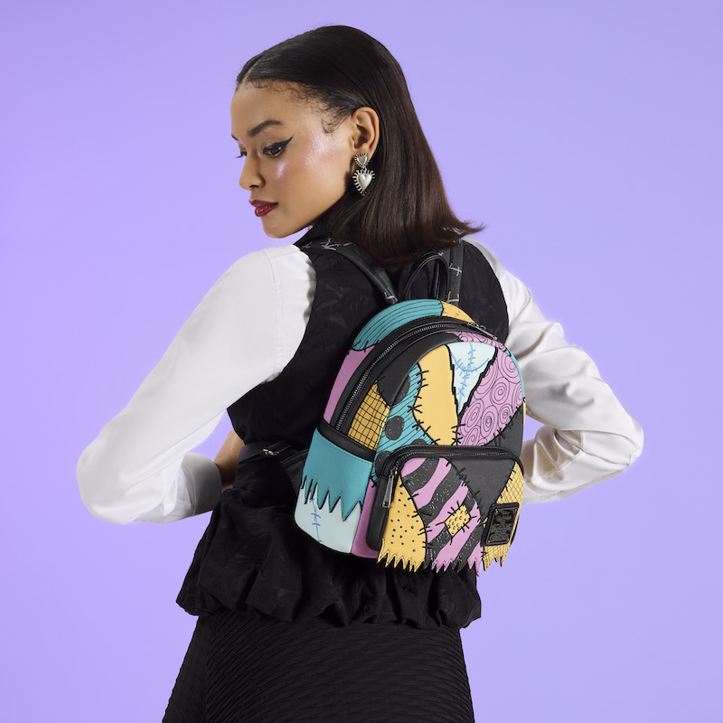 Woman facing away from camera wearing a white long-sleeve top and black vest. She wears the Loungefly Nightmare Before Christmas Sally mini backpack on her back. She stands in front of a purple background. 
